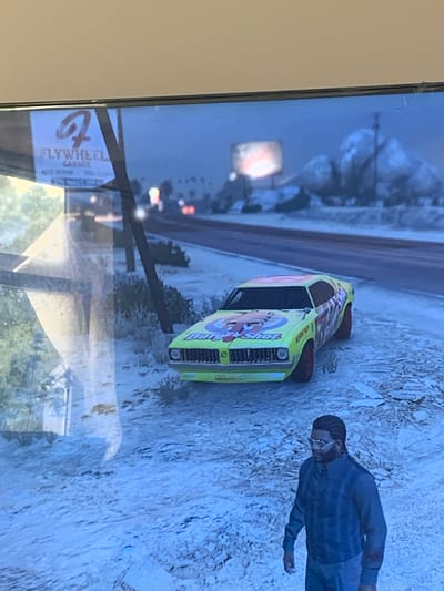 The image shows a video game scene with a yellow race car parked on a snowy road. A man wearing glasses and a blue jacket stands to the right of the car. In the background, there is a billboard and distant mountains. The environment appears to be an outdoor, possibly desert-like, setting with sparse vegetation and a cloudy sky. There are no elements in the image that suggest the location is Vejprty, Czechia.