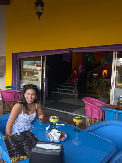 The image shows a young woman sitting at an outdoor table in front of a restaurant in Pozos, Costa Rica (coordinates: 9.951, -84.194). She is wearing a white floral dress and is smiling at the camera. The table is blue and has two glasses of margarita and a plate with a napkin and two small containers. There is a blue chair with a napkin dispenser on the right and a pink chair on the left. The restaurant has a large glass door with a purple frame and a yellow wall. The background is blurry and shows a staircase leading up to a brightly decorated space. The restaurant is decorated with colorful paintings on the wall and a sign that says "Sabores."