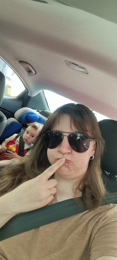 A woman wearing sunglasses and a brown shirt sits in the driver's seat of a vehicle. She is wearing a seatbelt and touching her upper lip with the index finger of her right hand. In the back seat, a young child sits in a car seat. The child wears a red and white shirt and holds a bottle. The interior of the car is visible, including the roof, seats, and dashboard elements. It is unclear whether the car is in Yongpyeong-myeon, South Korea, based solely on visual information in the image.