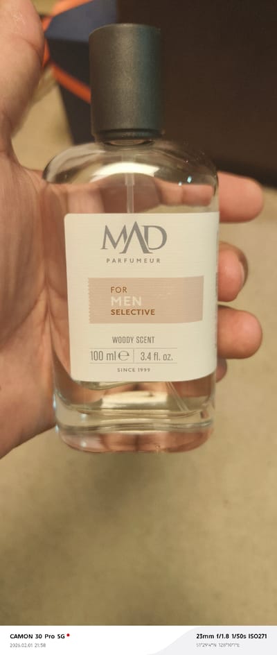 The image displays a clear glass spray bottle, identified as "MAD PARFUMEUR FOR MEN SELECTIVE," containing a clear liquid labeled "WOODY SCENT." The bottle, with a dark grey cap, indicates a volume of "100 ml e | 3.4 fl. oz." and states "SINCE 1999." An individual's hand, partially visible, holds the bottle vertically. The background is a soft-focus blend of dark brown and black tones, suggesting an indoor, dimly lit environment. Metadata at the bottom of the image indicates it was captured on 2026.02.01 at 21:58 by a CAMON 30 Pro 5G, with camera settings of 23mm f/1.8, 1/50s, ISO271. The GPS coordinates provided are 51°29'4"N 128°10'7"E, locating the scene within the Amur Oblast region of Russia, consistent with a Tecnimont camp.