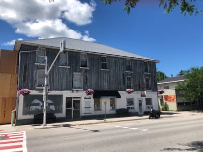 The image depicts the facade of a two-story building with a weathered, gray wooden exterior on the upper level and a white-painted storefront on the lower level. The building is situated in the city of Severn, Canada, likely in a commercial or mixed-use area, as suggested by the businesses visible on the lower level.

On the upper level, several arched windows are visible, some with white trim. The wooden siding appears old and weathered, with vertical planks. A light-colored metal roof tops the building. A pedestrian crossing sign with a silhouette of a person walking is mounted on a pole in front of the building.

The lower level features a white facade with multiple storefronts. One storefront, labeled "COLDWATER DENTURE CLINIC," displays information about services offered, including a phone number and website. Another storefront appears to be "TIFFANY'S OPEN TO LATE." Two large hanging flower baskets, filled with pink and purple blossoms, are suspended from the upper level near the storefronts. A black metal bench is placed on the sidewalk in front of the building. To the left of the storefronts, there's a large dark mural on the white wall, depicting what appears to be stylized wings or feathers in white paint.

A dark-colored motorcycle is parked on the street in front of the building. Across the street, a pedestrian crossing with red and white stripes is visible. The environment is a sunny day with a bright blue sky and scattered white clouds. Trees are present in the background to the right of the main building. The overall scene suggests a somewhat historic commercial street.