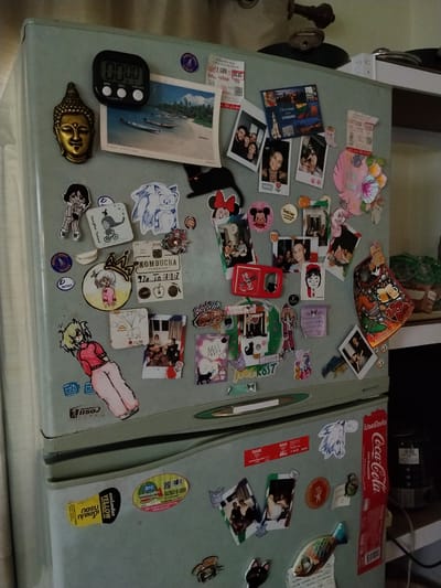 The image shows a close-up of a light blue refrigerator door covered with various magnets, stickers, and photos. On the top part of the door, a digital timer is visible, along with a small golden Buddha head figurine. Several printed photos are scattered across the surface, some appearing to be Polaroid-style snapshots with groups of people. Decorative stickers include cartoon characters like Mickey Mouse and Sonic the Hedgehog, as well as floral designs. A Coca-Cola branded item is prominently displayed on the right side of the lower half of the door. The refrigerator itself appears to be situated indoors, likely in a kitchen environment, with a glimpse of kitchen counters and appliances in the background. The context of Ban Patan, Thailand is not directly depicted or derivable from the visual elements of the image.