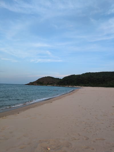 This is a wide-angle, outdoor photograph of a beach in Hoành Sơn Ward, Vietnam, taken from a low perspective. The sky is a pale blue with thin, wispy clouds. The ocean is a muted blue-green, with gentle waves lapping at the shore. The sand on the beach is light brown and has numerous footprints visible, indicating recent activity. In the distance, to the left, a large, tree-covered hill juts out into the sea. To the right, the beach extends further, meeting a dense, green forest that covers another hill. A few people can be seen swimming or wading in the water near the shoreline. There is a small piece of trash, possibly a wrapper, visible on the sand in the foreground. The lighting suggests it is either early morning or late afternoon.