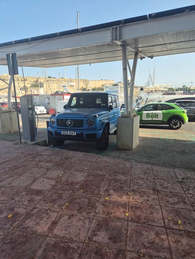 An eye-level, medium shot captures a blue Mercedes-Benz G-Wagon parked under a modern, flat-roofed structure with solar panels. A charging cable extends from a charging station to the car. To the right of the G-Wagon, a green electric vehicle with the "Bolt" logo is also parked. The vehicles are on a paved area. In the background, a harbor with sailboats and a fortified city wall is visible, suggesting the location is Ta' Xbiex, Malta. The sky is clear and blue, indicating fair weather. There are no people visible in the scene.