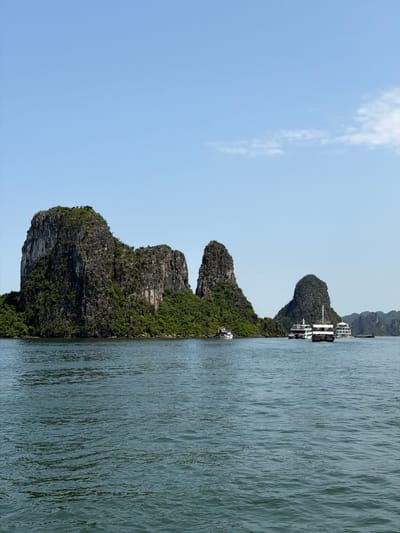 This landscape photograph, presented horizontally, captures a serene daytime view of a bay featuring distinctive limestone karst formations. Several islands, heavily covered in lush green vegetation, rise dramatically from the calm, blue-green water. The sky above is a clear, bright blue with a few wispy white clouds visible towards the left side of the frame.

In the middle distance, nestled between two of the larger islands, are two multi-deck boats, likely cruise ships or tour vessels, appearing to be anchored or docked close to the shore. One boat is prominently featured, showcasing its multi-level white structure. A smaller, single-deck boat is also visible closer to the shoreline of the island in the foreground. The water exhibits gentle ripples, reflecting the clear sky and surrounding scenery.

The scene suggests a tranquil moment in a picturesque natural setting. The bright, even lighting indicates it is likely morning or early afternoon, under fair weather conditions. The unique geological features strongly identify the location as Ha Long Bay, specifically consistent with the area around Tuan Chau Ward, Vietnam. No discernible text or individuals are visible in the image.