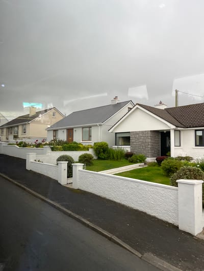 The image shows a row of detached houses on a street in Falcarragh, Ireland. The houses are of a similar style, featuring pitched roofs, white or light-colored walls, and gardens. The street in front of the houses is paved with asphalt and has a concrete curb. A white textured wall with square pillars borders the gardens of the houses. The sky is overcast, and there are reflections on what appears to be a window, possibly from a vehicle. The houses are well-maintained with manicured lawns and flowerbeds. One house has a prominent stone facade around its entrance. Telephone poles and wires are visible in the background.