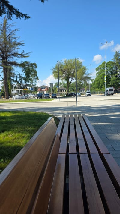 A low-angle shot captures a modern wooden park bench in the foreground, positioned on a paved walkway with a grassy area to the left. Beyond the bench, a street is visible, lined with parked cars and trees. In the background, a few buildings and streetlights complete the urban landscape under a clear blue sky. The scene appears to be in the city of Babyshira, Abkhazia.