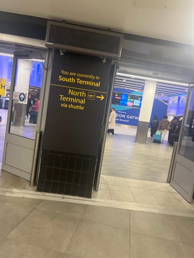 The image captures the interior of an airport terminal, specifically the South Terminal of London Gatwick Airport in Horley, United Kingdom. The scene depicts a brightly lit, modern environment with light-colored tiled flooring and blue accents on some information screens in the background.
The primary subject is a large, dark rectangular sign in the foreground, oriented vertically. It clearly states, "You are currently in South Terminal." Below this, an arrow pointing to the right, accompanied by an airplane icon, indicates the direction to "North Terminal via shuttle." Another digital display, partially visible to the left, shows text that appears to be "Do not enter" and "Departures Check in."
Several people are visible in the background, some with luggage, suggesting they are passengers either waiting, checking in, or transiting between terminals. Their attire suggests a cool or cold climate outside the terminal. A large information screen in the distance displays "Welcome to DON GATW" and "South Terminal," confirming the location. The overall atmosphere is typical of an airport, with individuals moving or waiting in a spacious, functional setting. The time of day cannot be determined precisely but the interior is well-illuminated.