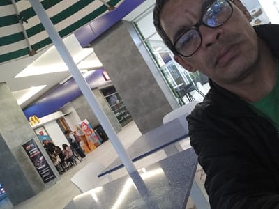 The image captures an indoor commercial dining area, identified as being in Tumbaco, Ecuador. A male subject, appearing mid-adult, with dark hair and wearing eyeglasses, a green shirt, and a dark jacket, is positioned in the foreground-right, looking directly towards the camera.
In the midground, several light blue speckled tables and at least one white chair are visible. A white vertical pole, possibly an umbrella support, extends from the lower frame towards the upper-left. Above it, a green and white striped umbrella is partially open.
The background features a McDonald's restaurant entrance, marked by its distinctive yellow "M" logo on a white fascia. To the left of McDonald's, a black vertical banner displays white text, including "VERSAL un mundo en tu bolsillo." To the right, a large promotional advertisement for soft-serve ice cream prominently features "50%" off. Several individuals are present in this background area: one adult female stands near the McDonald's counter, and three children are seated at a small table with stools.
The environment exhibits modern architectural elements, including textured grey walls and a ceiling featuring white and purple-blue panels with integrated lighting. Through a large window in the far background, additional indoor seating and tables are visible.