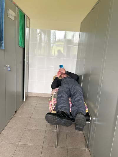 The image depicts a person lying on a narrow bench in a locker room or changing area. The person is wearing dark overalls and black boots and is looking at a blue smartphone. The bench is situated between rows of grey metal lockers on either side, with a tiled wall behind. A blue towel hangs from a locker on the left, and a green towel hangs from a locker to the right. The floor is tiled with square, greyish-brown tiles. The scene appears to be indoors, likely in a facility such as a sports center or industrial changing room. There is no discernible indication that this location is in the city of Подгорёна, Russia.