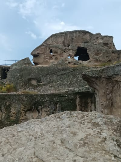 The image shows the Uplistsikhe cave complex in Georgia. The scene is an outdoor view of ancient rock-hewn structures. In the foreground, a large, rough, light-colored rock formation dominates the lower part of the frame. Behind it, a series of intricately carved caves and structures are visible within a larger, darker rock face. These structures feature numerous openings, including large arched entrances and smaller holes. The rock surfaces exhibit variations in color, from light brown to darker shades of gray and green, with patches of lichen or moss. Some vegetation, including dry grass and small bushes, is present on ledges and between the rock formations. A dark metal railing is visible on the upper left side, suggesting a pathway or safety barrier for visitors. The sky above is overcast with scattered clouds, indicating a daytime scene with diffused light. No people or animals are present in the image.