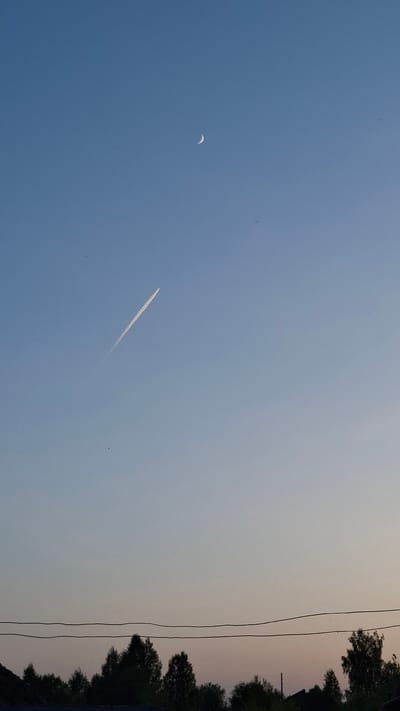 The image shows a sky in Шешурга, Russia. A crescent moon is visible in the upper left quadrant. A jet contrail is present in the middle of the sky. Trees and power lines are visible on the right edge of the frame. The sky transitions from blue to a light gray near the horizon. The image is oriented sideways.