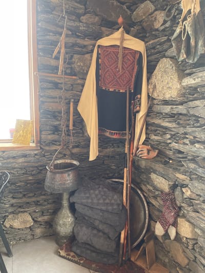 An interior corner, identified as being in Juta, Georgia, displays a collection of traditional objects against rough, dark grey stone walls. A rectangular window with a white frame on the left admits bright daylight, illuminating a golden, ornate object and a red-labeled item on its sill. Below the window, a hammered copper pot with a handle is suspended by a metal chain from a wooden support, positioned above a tall, slender metal vessel. To the right, a traditional black long-sleeved garment with intricate red, black, and white embroidered diamond patterns on the chest and lower sleeves hangs from a wooden hook; a plain light beige fabric is draped over its shoulders. Several wooden staffs lean against the wall in front of the garment, partially obscuring a large, round metal tray. A stack of approximately five dark grey, textured blankets or folded fabrics rests on a small patterned rug on the floor. On the right stone wall, a hanging brown, coarse textile object is visible, with a carved wooden hand resting on a stone protrusion below it. Further down, a patterned sock with red and white geometric designs hangs from the wall. No individuals are present in the scene.