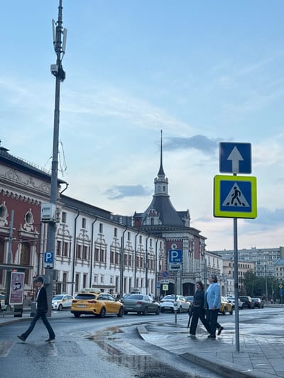 The image shows a street scene in an urban environment. In the foreground, a crosswalk sign with an arrow pointing left and a pedestrian crossing sign are visible. Two individuals are walking on the sidewalk, while parked cars and a yellow taxi line the street. The architecture in the background features a distinctive building with a pointed spire and detailed facade. A utility pole with mounted equipment is visible extending into the sky. The street appears wet. The location of Ukromne, Ukraine, is not verifiable based on image data.