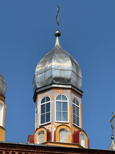 This is a close-up, low-angle, outdoor, daytime shot of the onion dome and upper facade of a religious building in Chudovo, Russia. The sky is a clear, bright blue. The main focus is on a large, silver-colored onion dome with a golden cross at its apex. The dome is constructed of segmented metal panels and shows signs of weathering and wear. Below the dome is a section of the building's facade painted in a faded yellow and orange color, with patches of peeling paint revealing a lighter colored underlayer. There are arched windows with white frames on this level, two of which are clearly visible. To the left, a portion of another, smaller silver onion dome is visible. The lower part of the image shows a dark red brick structure with a metallic roof. The overall impression is of an aging but still majestic religious structure against a clear sky. There are no people or animals visible in the frame.