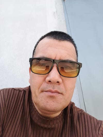 A man wearing sunglasses and a brown ribbed sweater is looking at the camera. He is standing in front of a white wall and a light blue metal surface. The sunglasses are black with orange lenses, and they reflect a street scene with a building. The man's hair is dark and short, and he has a fair complexion. The image is a close-up of his face and upper body. The scene is likely outdoors in a city setting.
