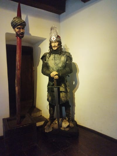 A wax figure of a medieval warrior stands in a dimly lit room with a white wall. The figure wears a dark, elaborate costume with armor and a feathered headdress. It holds a sword in its hands, which are crossed in front of its body. To the left of the warrior, a severed head is displayed on a red spike. The room appears to be part of a historical exhibit, possibly a museum or a themed attraction. The presence of the severed head suggests a theme related to historical battles or executions. The lighting is primarily from a source to the right, casting a shadow of the warrior on the wall. The floor is made of dark wood. This scene is likely located within Bran Castle, also known as Dracula's Castle, in the city of Predeluț, Romania, which is famous for its association with the legend of Count Dracula.