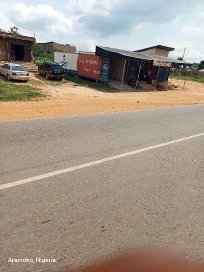 The image shows a paved road with a white dividing line in the center. There is a building on the right side of the road made from modified shipping containers. The container building has a sign on it that says "MAYFLOWER." The building to the left of the container building is partially obscured, but the sign on it indicates that it is a "Dangote Cement" dealer. There is a blue car parked in front of the building on the left. There is a second blue car parked behind the container building. There is a small, open-air structure in front of the container building. The image was taken in Okinni, Nigeria at coordinates: latitude: 7.832, longitude: 4.513. There is a small amount of vegetation on the sides of the road. The sky is partly cloudy. There are no people visible in the image.