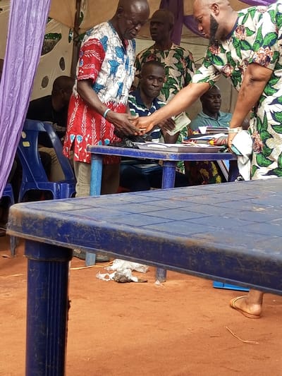 This image depicts a semi-outdoor gathering in Oko Town, Nigeria, under what appears to be a temporary fabric shelter or tent, distinguished by a purple fabric curtain on the left. The ground is reddish-brown earth, with some scattered litter visible.
Six adult males are present in the scene. In the central mid-ground, a bald man with a beard, wearing a white shirt with green and floral patterns, is engaged in an exchange with an older, bald man with dark skin. The man in the floral shirt extends his right hand, seemingly transferring a small black plastic bag to the older man, who receives it with both hands. The older man wears a red, white, and blue patterned short-sleeved top over light-colored trousers and has a light blue wristband on his left wrist. The man in the floral shirt also holds several Nigerian Naira banknotes in his right hand, along with a piece of white fabric or paper in his left.
Behind this interaction, a man with dark skin, wearing a green and white striped polo shirt, is seated at a blue plastic table, looking downwards. To his right, another bald man, dressed in a green and white patterned shirt similar to the floral one, stands and observes the transaction. In the background, partially visible, are two more men: one in a dark shirt, seated on a blue plastic chair on the left, and another in a light green shirt, seated at a table on the right.
A large, weathered blue plastic table dominates the immediate foreground, partially obscuring the lower half of the subjects and the ground. The scene captures a focused interaction or transaction occurring among the individuals.