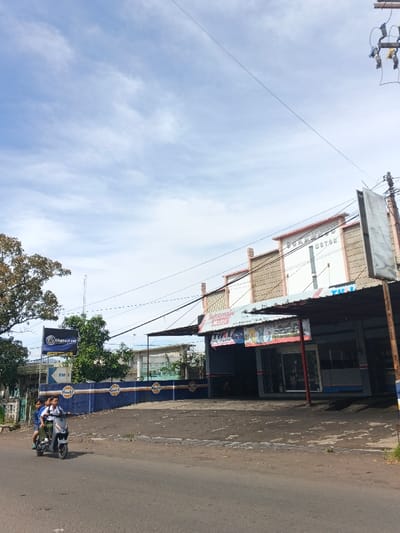 A daytime street scene presents a motorcycle carrying three people, driving on a road in front of a building. The building's facade is adorned with a blue and orange stripe design and Gulf Oil logos. "Sukamaju Motor" signage is visible above the storefront. Utility poles with wires are on the right, and some trees are visible on the left. The sky is partly cloudy.