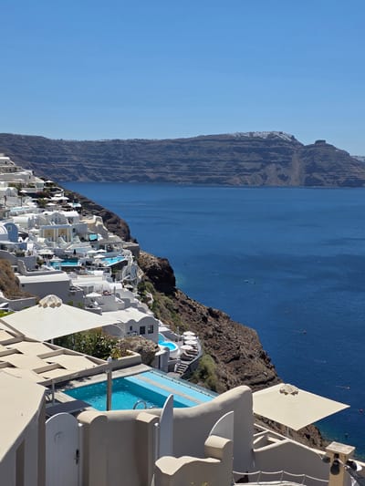 This is a landscape photograph taken from a high vantage point overlooking a coastal village. The scene is dominated by the deep blue of the sea and the clear, bright sky, suggesting a sunny day. The village is built on a steep cliff face, with a dense cluster of white, cubic buildings cascading down towards the water. Many of these buildings feature rounded edges and are adorned with blue accents, a characteristic architectural style often associated with Greek islands.
Prominently visible are several swimming pools, their turquoise water contrasting with the white structures. The architecture is modern but incorporates traditional elements, with balconies, terraces, and open spaces. The sea is calm with a few small boats visible in the distance, suggesting it is a peaceful day. The rugged, rocky cliff face on which the village is built provides a dramatic backdrop.
The perspective suggests the photo was taken from a balcony or higher level of one of the buildings, looking down and across the village and out to the sea. The time of day appears to be late morning or early afternoon, given the bright sunlight casting sharp shadows. The overall impression is one of luxury and tranquility, typical of a holiday resort in a scenic coastal location. The specific location of Ia Municipal Unit, Greece, points to the iconic island of Santorini, known for its white villages perched on the caldera cliffs. There is no visible text in the image.