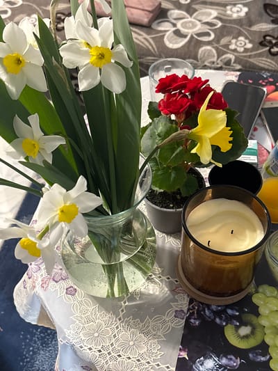 This is a close-up shot of a still life arrangement. The primary focus is on a vase of white daffodils with yellow centers, alongside other plants with red and yellow flowers. A brown glass candle sits nearby. The arrangement is placed on a table covered with a patterned tablecloth and a blue surface. Other objects visible include a jar of orange juice, a cell phone, and a box of cigarettes, among other items. The location is not discernible from this image. Novo Panicharevo, Bulgaria is not evident.