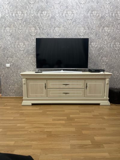 The image shows a room interior with a gray patterned wallpaper, a black flat-screen television mounted on the wall, and a light-colored cabinet with two doors on the top and two doors on the bottom standing on a wooden floor. An unidentified black object is visible above the cabinet and in the bottom right corner of the image. There is no indication of the image's location as Akhalkalaki, Georgia. No people, actions, or interactions are depicted.