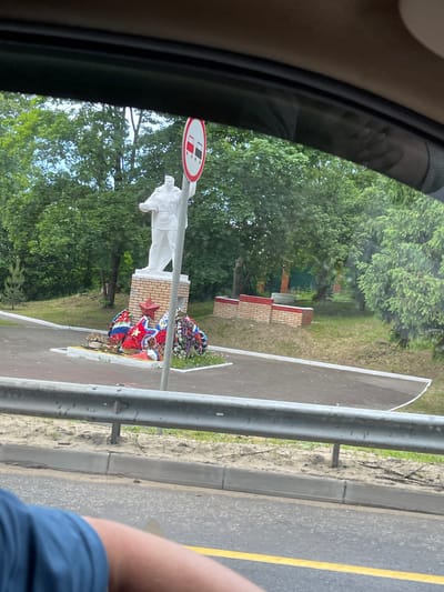 Captured from within a vehicle, the image reveals a monument commemorating soldiers in Semyonovskoye, Russia. The monument comprises a white statue of a soldier set on a brick pedestal. Wreaths adorn the base. A sign is visible above the statue. Trees form the backdrop. A road with a yellow line is seen along with a metal guardrail. An arm wearing a blue shirt is visible in the lower right corner of the frame. The scene is viewed through the car's window frame.
