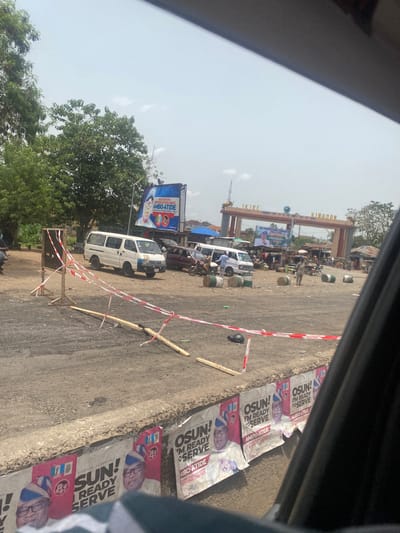 The image depicts a street scene in Ikire, Nigeria. There is a large sign archway that says “IKIRE KINGDON” (Kingdom). Alongside the road, there are several posters promoting candidates with the name “AMBO-ATIDE” and slogans such as “SUN! I’M READY TO SERVE”. There are parked vehicles, including a white van, along the side of the road. There are red and white barricade tapes present. Trees and buildings are visible in the background. People can be seen in the background behind the sign archway.