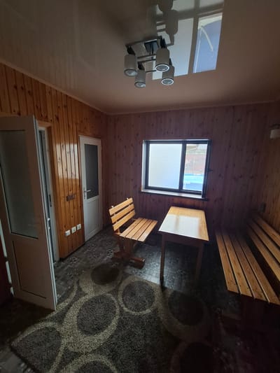 The image depicts the interior of a wooden room, likely a rustic cabin or sauna. The walls and ceiling are paneled with light-colored wood, creating a warm and natural ambiance. Sunlight streams in from a large window, casting geometric shadows on the floor.

In the foreground, a light-colored wall dominates the left side of the frame. Mounted on this wall is a modern light fixture with three cylindrical lamps, their white shades reflecting the ambient light. Adjacent to this, a large white framed window or door is partially visible, suggesting an exit or another section of the building.

The center of the image features a square-shaped wooden table and two matching wooden benches. The table has a slightly reflective surface, hinting at a recent cleaning or the type of wood used. The benches are simple, with slatted seats and backs, designed for outdoor or casual seating. Beyond the table and benches, a small, square window with frosted glass is set into the wooden wall.

The floor is a mosaic of different textures and patterns. In the area around the table, the floor appears to be paved with small, irregularly shaped stones or tiles, creating a somewhat uneven surface. Further back, a large, circular patterned rug is visible, its dark color contrasting with the lighter wooden elements.

The overall scene suggests a tranquil and natural setting. The time of day appears to be daytime, given the sunlight, but the precise hour is not discernible. There are no people or animals visible in the image, and no text is present. The setting feels calm and possibly private, fitting for a retreat or a leisure space. The wooden construction and the presence of a rug suggest an indoor space designed for relaxation or communal gathering.