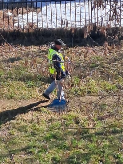 The image depicts an outdoor scene in Yegoryevsk, Russia. A single individual is walking from left to right across a ground covered with sparse green grass, numerous dried brown leaves, and twigs. The person wears a dark baseball cap, a dark long-sleeved garment underneath a high-visibility lime green/yellow reflective safety vest, light-colored pants, and dark footwear. They are holding a blue-headed leaf rake by its handle, with the rake head positioned near the ground. A distinct shadow of the individual and the rake extends to the left on the terrain. In the upper background, a dark metal fence with vertical bars is visible, behind which appear patches of white material, consistent with snow or ice, interspersed with brown vegetation.