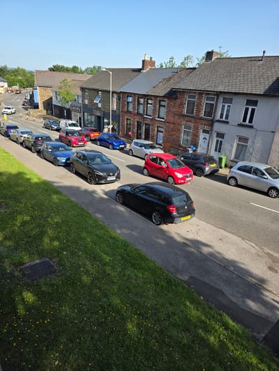 This is a vertically oriented, eye-level, outdoor photograph taken on a sunny day, likely in the afternoon, in Cwmbrân, United Kingdom. The scene depicts a residential street lined with terraced houses and parked cars. The houses are a mix of brick and rendered exteriors, with multiple stories and visible windows. The street is paved, with a curb separating it from a grassy area on the right side of the frame, which is dotted with dandelions.
A variety of cars are parked along the street, including a silver hatchback, a dark grey hatchback, a red hatchback, and a black hatchback. Further down the street, more cars are visible, including a blue hatchback, a white van, and other vehicles. The parked cars suggest a typical residential setting where residents are using their vehicles. There are no people clearly visible in the foreground, though some figures might be present in the windows of the houses or further down the street, obscured by distance or angle.
The time of day is suggested by the bright sunlight and the shadows cast on the pavement and grass, indicating a clear, sunny day. The environment appears to be a typical urban or suburban residential area in the UK, characterized by the architecture of the houses and the presence of parked cars. The overall impression is one of a quiet, ordinary day in a residential neighborhood. No prominent text is visible on the buildings or any of the objects within the frame, apart from what may be on the car license plates.