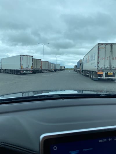 The image shows a parking lot filled with semi-trailer trucks under a cloudy sky. The perspective is from inside a vehicle, looking out at the parked trucks. In the foreground, the dashboard and a digital display are visible, showing a temperature of 26.0°C and some icons. The parking lot appears to be a rest stop or service area, with multiple lanes and rows of trucks. Some trucks have company logos and markings, including Chinese characters on one of the trailers. The scene suggests a commercial transportation hub. The location is identified as Kliuchiki, Russia, although there are no specific visual cues in the image to confirm this without external information.