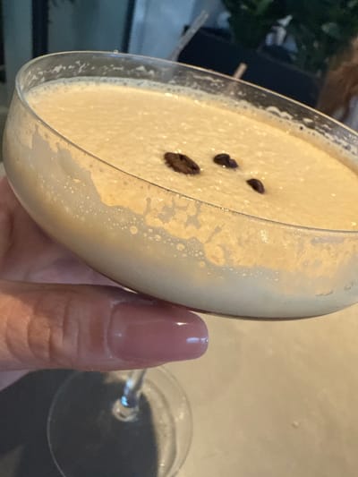 A close-up shot depicts a person holding an espresso martini in a cocktail glass, garnished with three coffee beans. The beverage has a creamy, foamy appearance. The person's hand with a nail painted mauve color holds the glass at an angle. The scene is indoors, possibly in a bar or restaurant in Oak Brook, United States of America. The background is blurred, with some plants and a dark object visible. Another empty cocktail glass can be seen to the right of the main subject.
