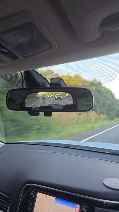 The image shows a view from inside a vehicle traveling on a highway. A rear-view mirror reflects cars ahead on the road, which is lined with trees. The dashboard of the car is visible in the foreground, with a GPS screen showing a map and the time 6:46. The environment appears to be a rural area, possibly near Cherwell District, United Kingdom, given the commonality of such highways and vegetation in that region. The lighting suggests it is late afternoon or early evening. There are no people visible in the frame, and the action is simply driving.