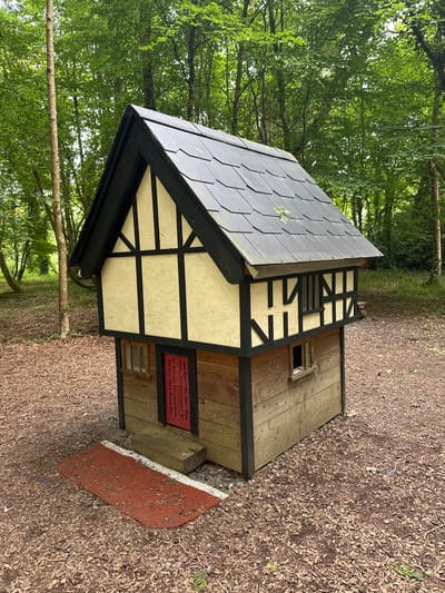 A small, Tudor-style playhouse sits on a woodland floor covered in wood chips and fallen leaves. The playhouse has a dark gray shingled roof, black trim, and cream-colored walls with black timber framing details. It features a red door, two small windows on the front, and a smaller window on the right side. A wooden step leads to the red door, and a red doormat with white edging is placed in front of it. The playhouse is surrounded by trees with green foliage. The scene is likely located in the grounds of Castlecomer Estate in County Kilkenny, Ireland, known for its woodland walks and attractions.
