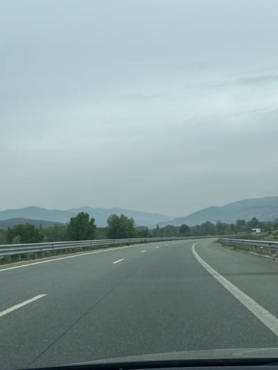 The image depicts a highway cutting through a mountainous landscape, possibly in or near Strumyani, Bulgaria. The road is grey asphalt, marked with white lines. A metal guardrail runs along the right side of the highway. The environment shows a hazy, overcast sky. Mountains are partially obscured by the haze, with trees visible along the roadside. There are no people visible in the image.
