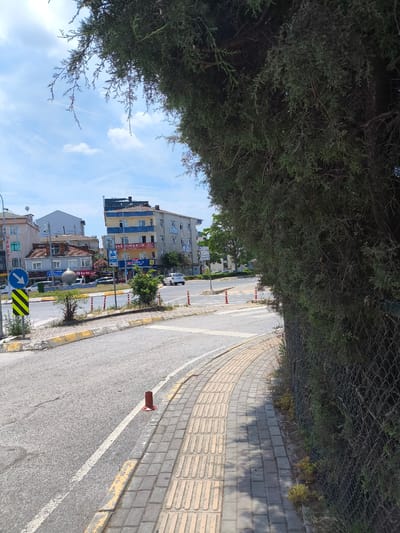 A street scene in Tuzla, Turkey, captured on a sunny day with scattered clouds. The foreground is dominated by a sidewalk paved with tactile paving bricks, a common feature for visually impaired pedestrians. Adjacent to the sidewalk is a chain-link fence partially covered by dense green foliage from a large tree, whose branches overhang the scene from the right. The road curves to the left, marked with yellow and white lines. Orange traffic cones are placed along the road.

In the background, several multi-story buildings line the street. One building, taller than the others, has a facade with blue and yellow sections. Signs on the buildings indicate commercial establishments, with one prominent sign reading "JAS MIMARLIK" in large letters. Other signs are visible but difficult to decipher. A white car is parked on the street, and another is driving away in the distance. A blue and white directional sign with an arrow pointing left is mounted on a pole. A circular, sphere-like sculpture is visible in the middle of a roundabout in the distance. The overall impression is a typical urban street environment in Turkey during daylight hours.