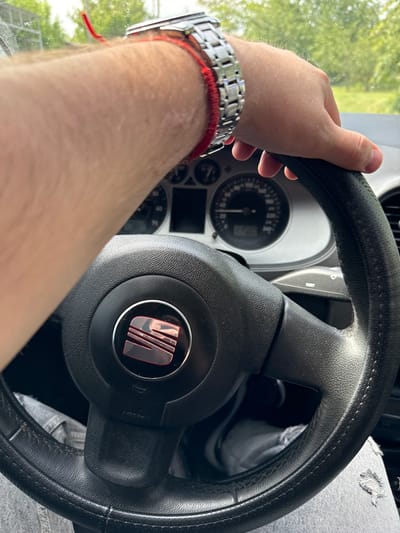 A first-person perspective image captures a person's right hand and forearm gripping a black leather-wrapped steering wheel in a car during the daytime. The hand, with visible arm hair, wears a metallic link wrist watch and a red string bracelet. The steering wheel, likely showing signs of use with slight wear on its textured surface and outer rim, features a red and metallic SEAT logo centrally on its hub. The word "AIRBAG" is visible on the right spoke of the steering wheel.
Behind the steering wheel, the car's instrument cluster is partially visible, showing a speedometer with numerical markings (e.g., 20, 40, 60, 70, 80) and a needle pointing towards a low speed or zero, suggesting the car is stationary or moving slowly. Through the windshield, outside the car, bright green foliage from trees and a bright sky indicate a clear, sunny day. The person's leg, wearing distressed light-colored denim jeans, is partially visible in the lower right corner of the frame. The overall scene depicts someone inside a vehicle, possibly in Donets, Ukraine, preparing to drive or paused during a journey.