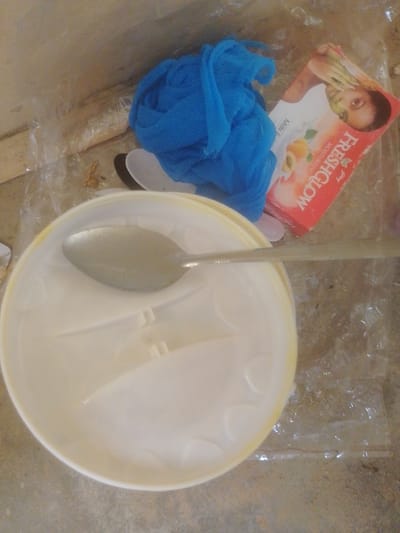In Majemu, Nigeria, an overhead shot captures a collection of items on a surface that appears to be a floor with visible debris. A white plastic tub, partially filled with a light-colored substance, dominates the lower portion of the frame. A silver spoon rests inside the tub, angled diagonally. To the upper right, a small red box, likely packaging for a cosmetic product, is visible, with the words "Fresh & Glow" and an image of a woman's face prominently displayed. Beside the tub, a crumpled blue net material is present. The surrounding environment shows signs of disarray, with scattered plastic sheeting and small pieces of debris, suggesting an unkempt or storage area.