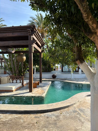 The image shows a luxurious outdoor space with a swimming pool and a wooden pergola. The pool is kidney-shaped and filled with turquoise water. It is surrounded by a concrete patio and a light-colored stone border. To the left of the pool, under the pergola, is a daybed with white cushions. A wooden bench is also visible next to the daybed. The pergola itself is made of dark wood and features intricate carvings. Several potted plants, including palms and other tropical foliage, are scattered around the area. A large, round, textured lantern hangs from the pergola. In the background, a white building with large windows and doors can be seen. The overall impression is one of a serene and upscale vacation setting. The scene is set in Sant Joan de Labritja, Spain.