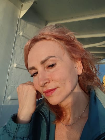 A woman with reddish-pink hair is captured in a close-up selfie, her face angled towards the viewer. She rests her chin on her right hand, which is positioned near her cheek. Her eyes are cast downwards, and her lips are slightly parted, revealing a hint of a smile. She is wearing a teal-colored jacket and a delicate silver necklace. The background appears to be a structure with painted white and light blue walls and a white ceiling, possibly on a boat or an outdoor area with overhead cover. The lighting suggests it is either sunrise or sunset, casting a warm glow on her skin. No specific details about Gorushka, Russia are discernible from the image.
