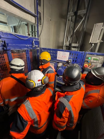 Five construction workers in full personal protective equipment are depicted in a confined industrial environment, likely a vertical shaft or tunnel under construction, in Aulnay-sous-Bois, France. All individuals wear high-visibility orange and dark blue jackets with reflective stripes, hard hats (white, yellow, black, silver/grey), and black neck gaiters; some also wear ear protection muffs. They are clustered around a blue industrial hoist or access platform control unit. An individual on the far right is observed interacting with the control panel, which displays green indicator lights, a screen, and various buttons. A label on the machinery indicates a "CMU: 1000kg," signifying a maximum useful load of 1000 kilograms. The background reveals raw concrete walls, exposed metal structural elements, and a vertical mast likely associated with a lifting apparatus.