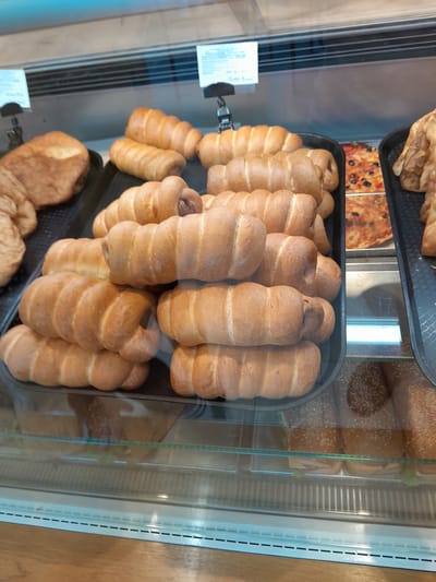 In Cimișlia, Moldova, a glass display case at a bakery showcases an assortment of baked goods. The primary focus is on a tray of golden-brown, sausage-filled pastries, with multiple rows stacked upon each other. To the left of the pastries, a large, irregularly shaped bread roll is visible. To the right, a slice of pizza with red sauce, black olives, and what appear to be pieces of meat is displayed in a black tray. Further to the right, another tray holds what looks like fried pastries. Below the main display, on a lower shelf, sandwiches with sesame seed buns and visible fillings are arranged. The lighting in the bakery highlights the textures and colors of the food items.