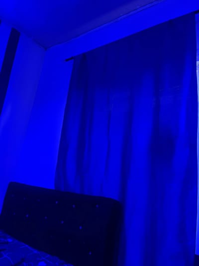 This media file captures an interior scene illuminated by vibrant blue light, creating a somewhat abstract and moody atmosphere. The primary subjects are architectural elements and what appears to be a piece of furniture or a decorative panel.

In the upper portion of the image, a large, dark blue surface with visible creases and folds dominates the frame, suggesting fabric or a similarly textured material. Below this, a diagonal plane of the same blue material is seen, angled downwards. To the right, a black, rectangular object is visible, possibly a shelf or a panel, which is studded with numerous small, circular, light-colored flecks that seem to reflect the ambient blue light. A portion of what looks like a patterned surface, also in shades of blue and black, can be seen on the far right edge. The left side of the image shows a corner formed by two blue-lit walls, with a thin vertical line suggesting a seam or an edge.

The scene is characterized by the intense blue lighting, which casts deep shadows and highlights the textures of the surfaces. There is no visible indication of time of day or weather, as the interior setting is entirely controlled by artificial light. The location context provided is Pantinapu, Nigeria, but there are no specific visual cues within the image that confirm this location. The overall mood is enigmatic and perhaps a little futuristic due to the pervasive blue glow. No people are visible, and no text is present in the image. The activity taking place is not discernible; it appears to be a still, captured moment of an interior space.