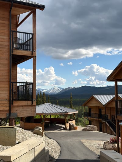 The image depicts a view from a property in Denali National Park and Preserve, United States, with a focus on wooden lodge-style buildings set against a backdrop of mountains and a forest. In the foreground, a paved road curves through a gravel-covered area. To the left, a multi-story wooden building with balconies and black railings is visible. A covered pavilion with a metal roof, picnic tables, and seating is centrally located. To the right, another similar wooden building with multiple levels, balconies, and exterior staircases is present. The background features a dense forest of evergreen trees leading to a range of mountains, some of which are capped with snow. The sky is partly cloudy with dramatic, large grey and white cumulus clouds against a blue sky. There are no people visible in the image. The scene suggests a lodge or resort accommodation in a mountainous, natural setting.