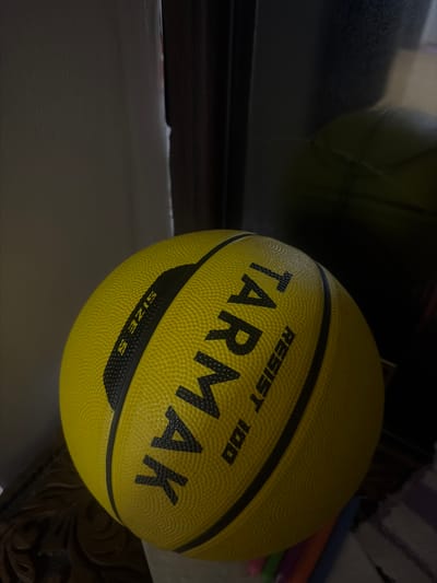 A yellow basketball with black lettering is positioned on a wooden surface. The lettering on the basketball reads "TARMAK" and "RESIST 100". The word "SIZE S" is also visible in black lettering. The basketball is casting a shadow on the surface it rests on. The background is dark, with a reflection of the basketball and the surrounding environment visible in what appears to be a mirror or reflective surface. The scene is dimly lit, with the light source seemingly coming from the left. No people are present in the image. There is no indication of the city of Khyrdalan, Azerbaijan.