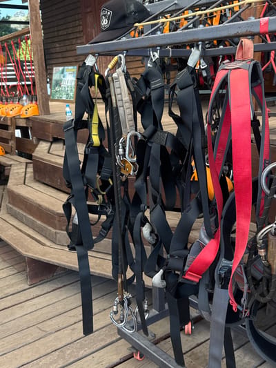 A metal rack holds several harnesses, mostly black with some red and yellow, for a high ropes course or ziplining. Metal clips and other hardware are attached to the harnesses. In the background, a wooden structure is visible, along with a glimpse of a red rope bridge. Some helmets are visible at the lower edge of the image. A black baseball cap is on the rack.