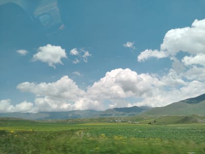 The image depicts a rural landscape under a bright blue sky dotted with fluffy white clouds. In the foreground, a vibrant green field stretches out, speckled with yellow wildflowers. A blurry streak of green and yellow suggests movement, likely indicating the photo was taken from a moving vehicle.

In the mid-ground, rolling hills covered in green vegetation rise gently. Nestled among these hills is a small cluster of buildings, appearing to be a village. Some of the buildings are white, while others have darker roofs. A few scattered trees are also visible.

The background is dominated by a range of majestic mountains, their peaks softened by distance and possibly a light haze. The overall impression is one of a serene, pastoral scene in the Armenian countryside. While not explicitly labeled as Mirak, Armenia, the visual elements are consistent with the geography of such a location. No people or specific actions are discernible in the image.