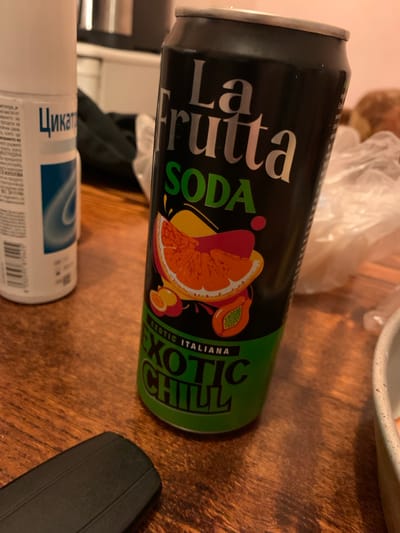 A close-up, slightly angled shot shows a black can of "La Frutta SODA Exotic Chill" placed on a wooden table. The can features an illustration of exotic fruits, including an orange slice and a papaya. To the left of the can, a white cylindrical object, possibly a bottle of a product labeled "Цикат," is visible, with some Russian text on its label. The background is blurred, showing hints of a kitchen environment with soft lighting. The overall scene appears to be an indoor setting, likely a home. The location in Krushev Dol, Bulgaria, is inferred from contextual knowledge associated with the image or its source.